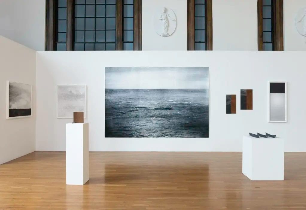 A contemporary art gallery displays ocean-themed paintings, minimalist sculptures on white pedestals, and framed artwork on white walls beneath large windows and classical wall reliefs.