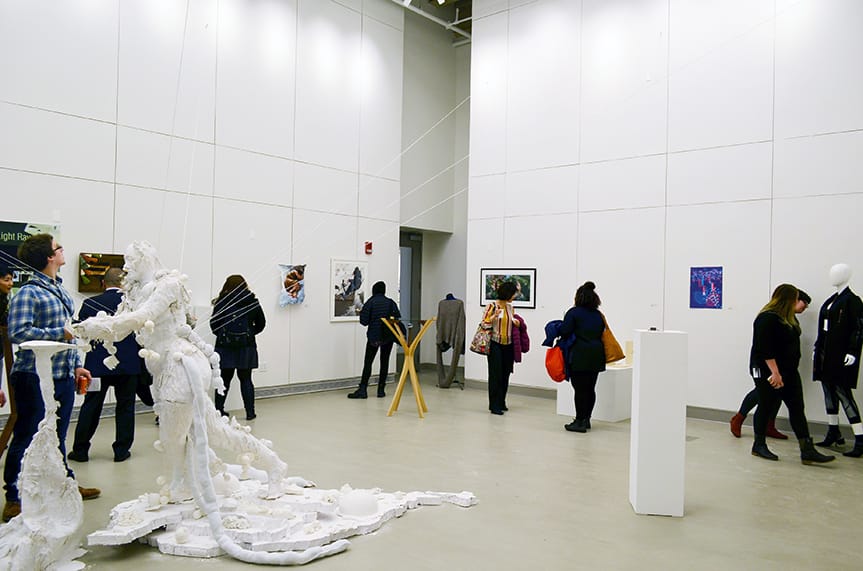 In a modern gallery, people view artwork in a room with white walls, where various pieces, including sculptures and framed art, are displayed. Amidst the casual visitors interacting with exhibits is Marion, enjoying the ambiance before attending a nearby glass blowing demonstration.