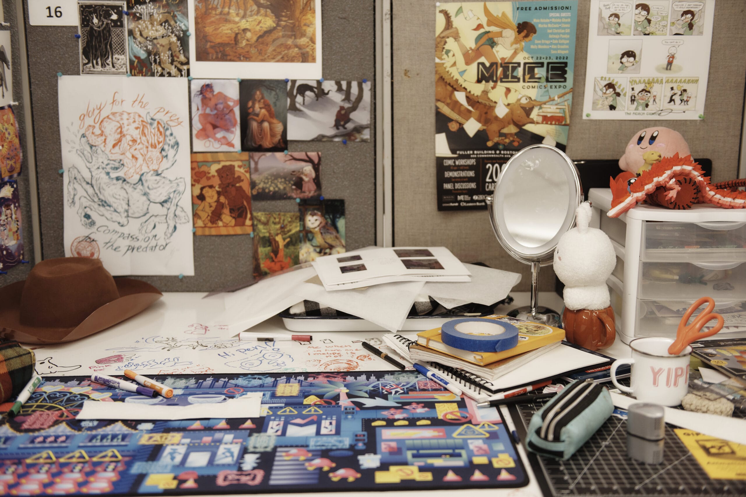 A cluttered artists workspace bursts with comic arts as colorful drawings, posters, and sketches are pinned to the wall. The desk holds markers, notebooks, a lamp, a hat, and a mug with Yip, alongside various visual storytelling tools. A small mirror and plush toy complete the scene.