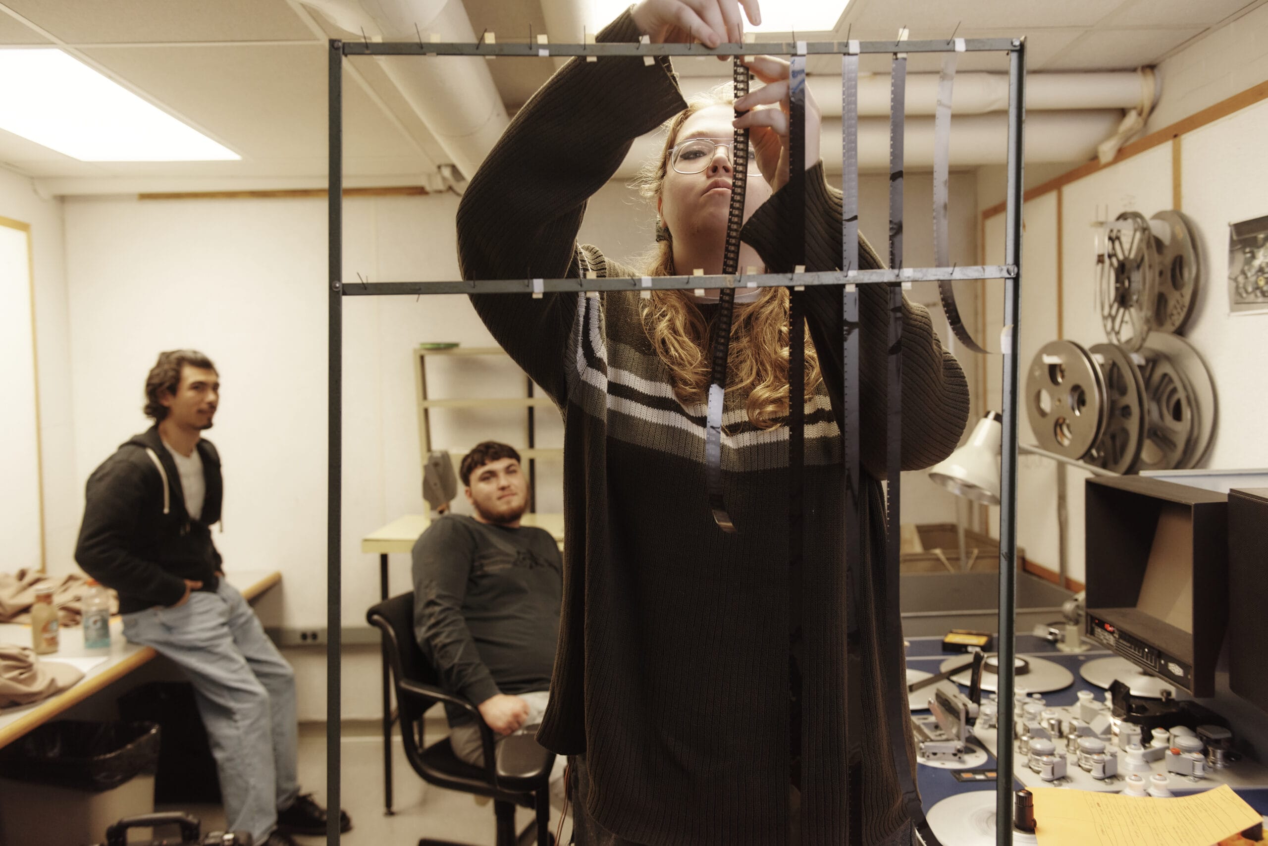 A person adjusts film strips on a rack in a room lined with state-of-the-art equipment, reflecting the high standards of the best film/video program. Two others sit in the background, one observing attentively from a chair amidst film reels and a desk with a cutting board.