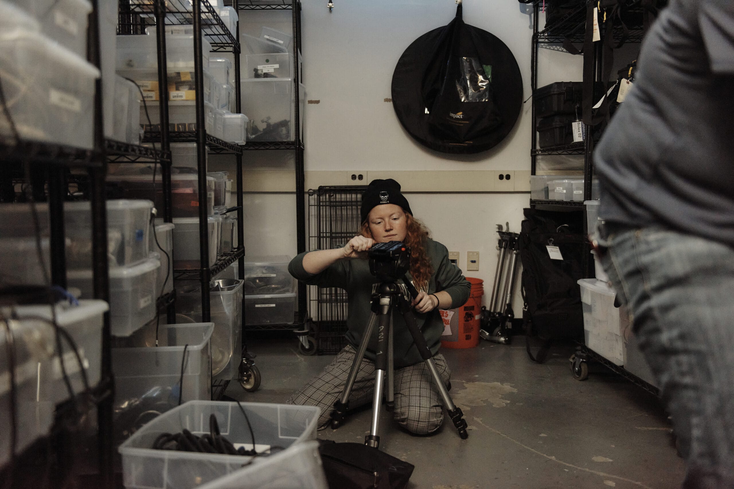 In a storage room brimming with plastic bins, a long-haired individual wearing plaid pants and a beanie crouches on the floor. Theyre setting up a camera on a tripod, likely capturing scenes for their major in animation project.