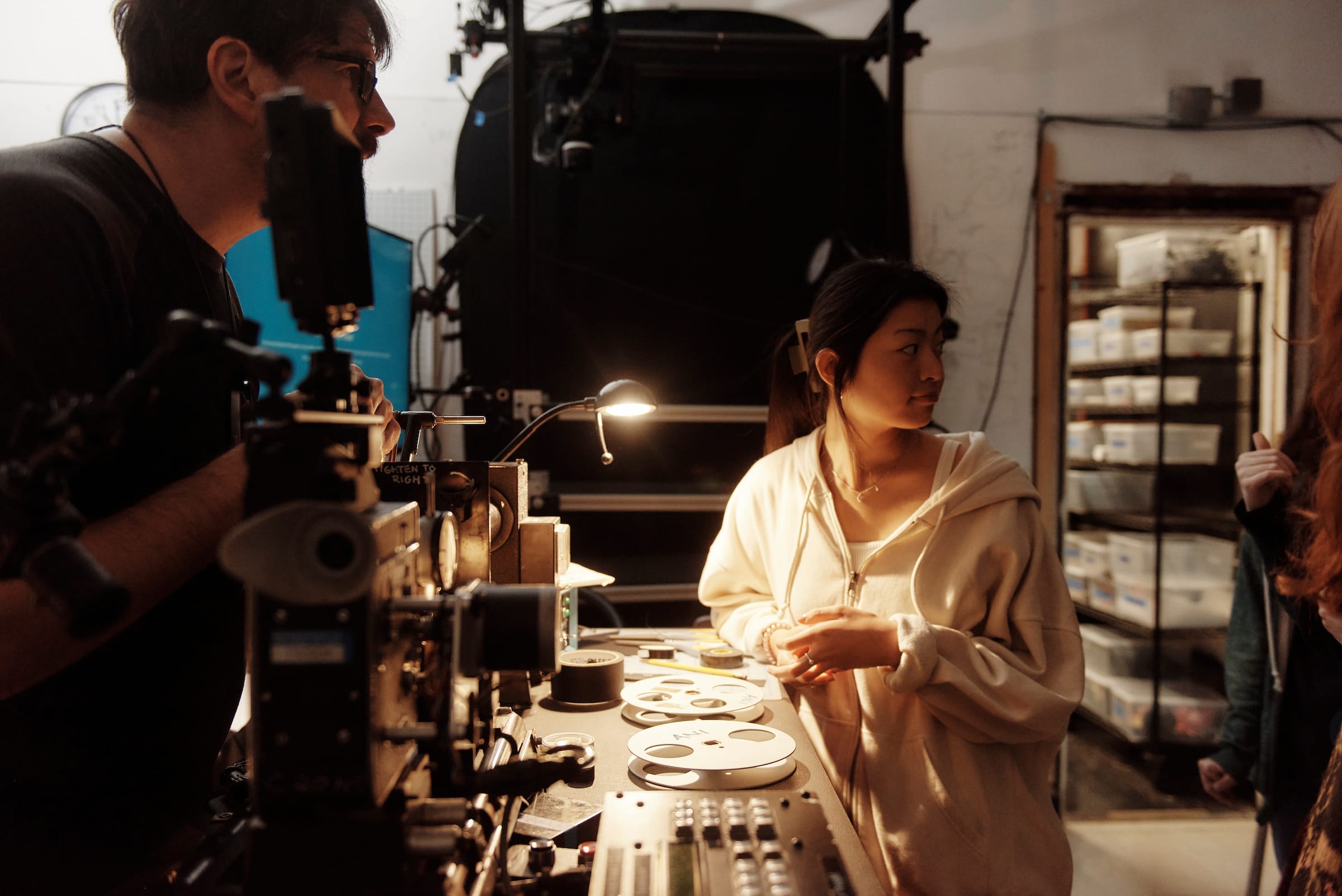 A woman in a light hoodie stands by a film editing machine, surrounded by film reels, while a man speaks nearby in a cluttered animation workspace with shelves of labeled boxes—reflecting the creative energy of a BFA studio.