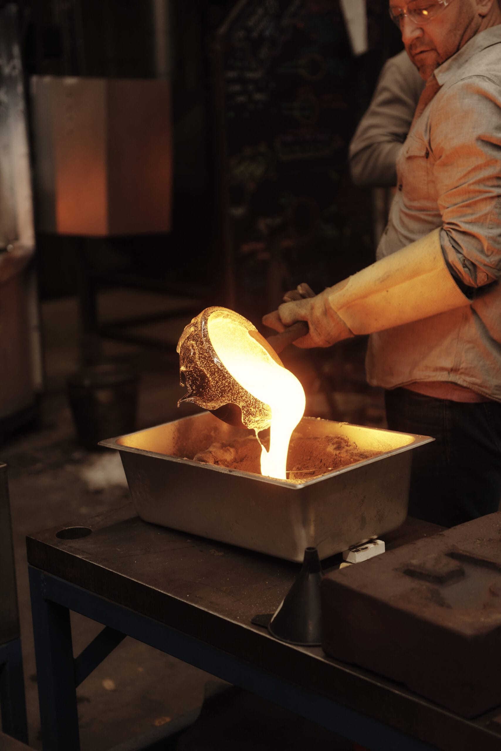 A worker wearing protective gear pours molten metal from a crucible into a mold on a workshop table. The glowing liquid casts light, illuminating the dark industrial environment.