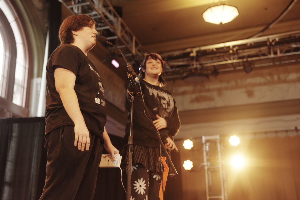Two people stand on stage, immersed in the performing arts, smiling as they hold microphones. Dressed casually under warm lighting with a decorated backdrop, one wears a black T-shirt and pants; the other, a black shirt paired with a floral skirt.