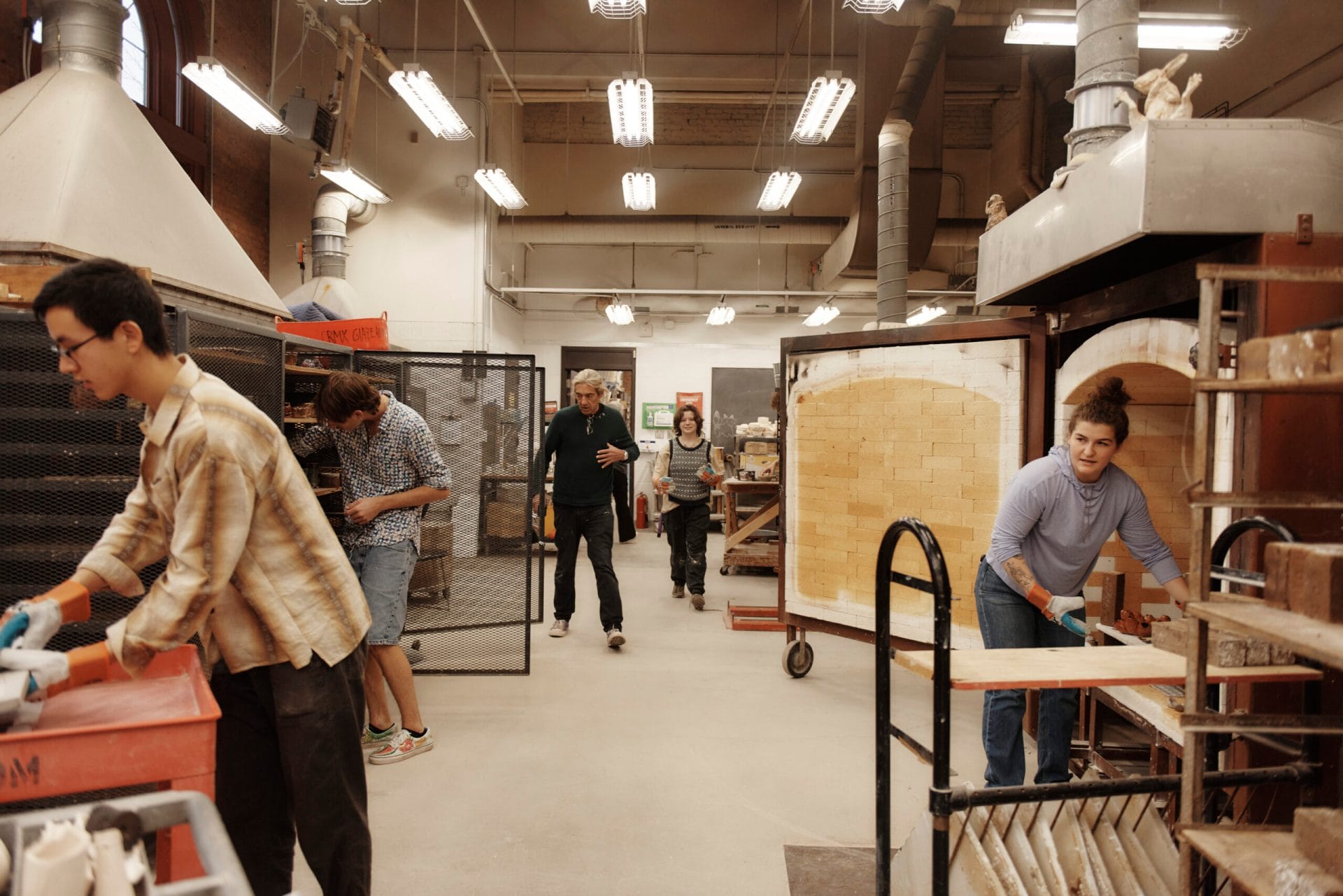 In this vibrant pottery studio, where many hone their craft, one person carefully places a piece into the kiln as others expertly mold clay and handle tools. Shelves of ceramics and equipment fill the backdrop—a testament to the artistry often fostered in a BFA program.