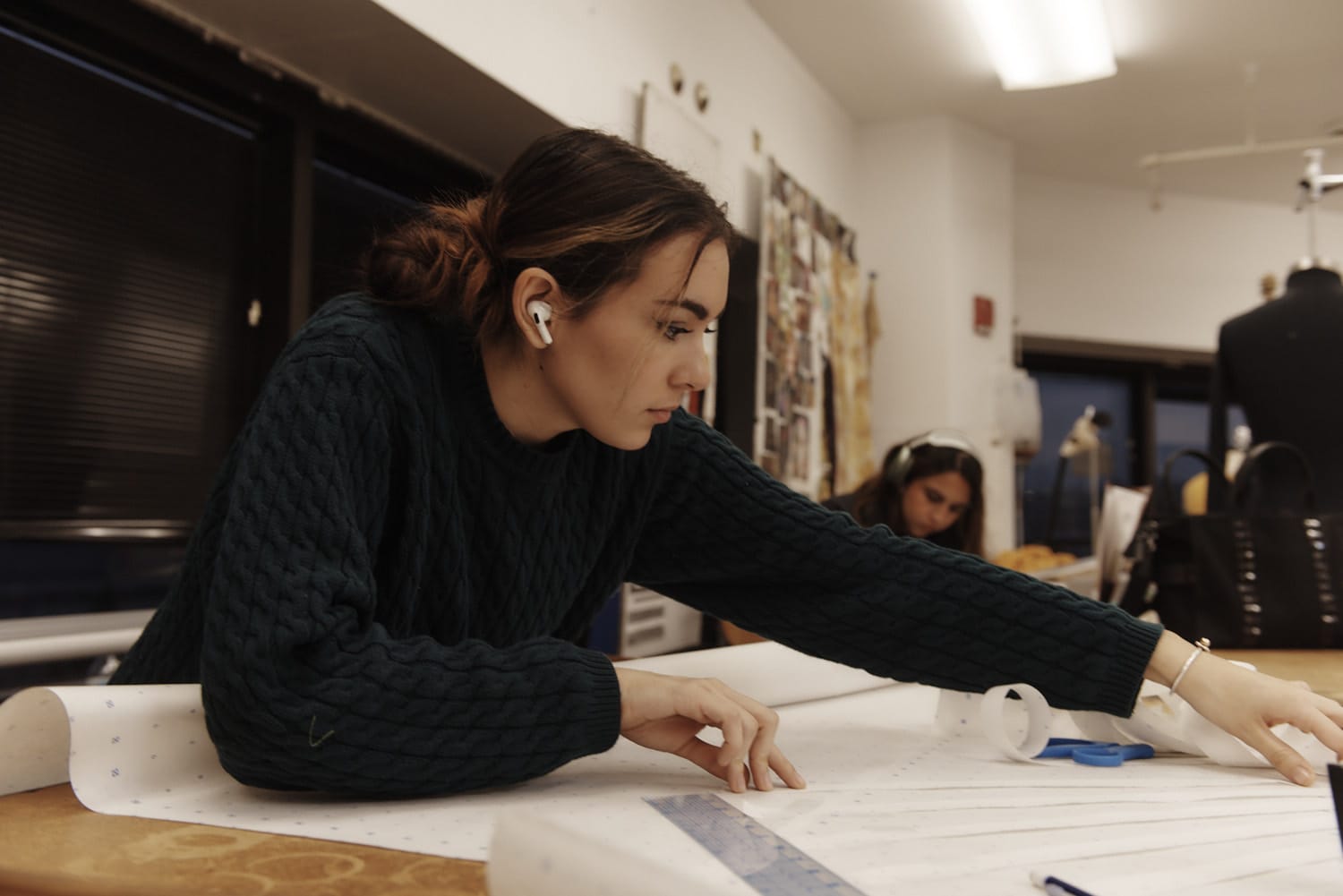 A person in a green sweater and earbuds is focused on measuring fabric with a ruler on their work table, showcasing the dedication typical of MassArt Fashion Design. In the background, another individual, wearing headphones, works amidst well-lit surroundings and scattered sewing tools.