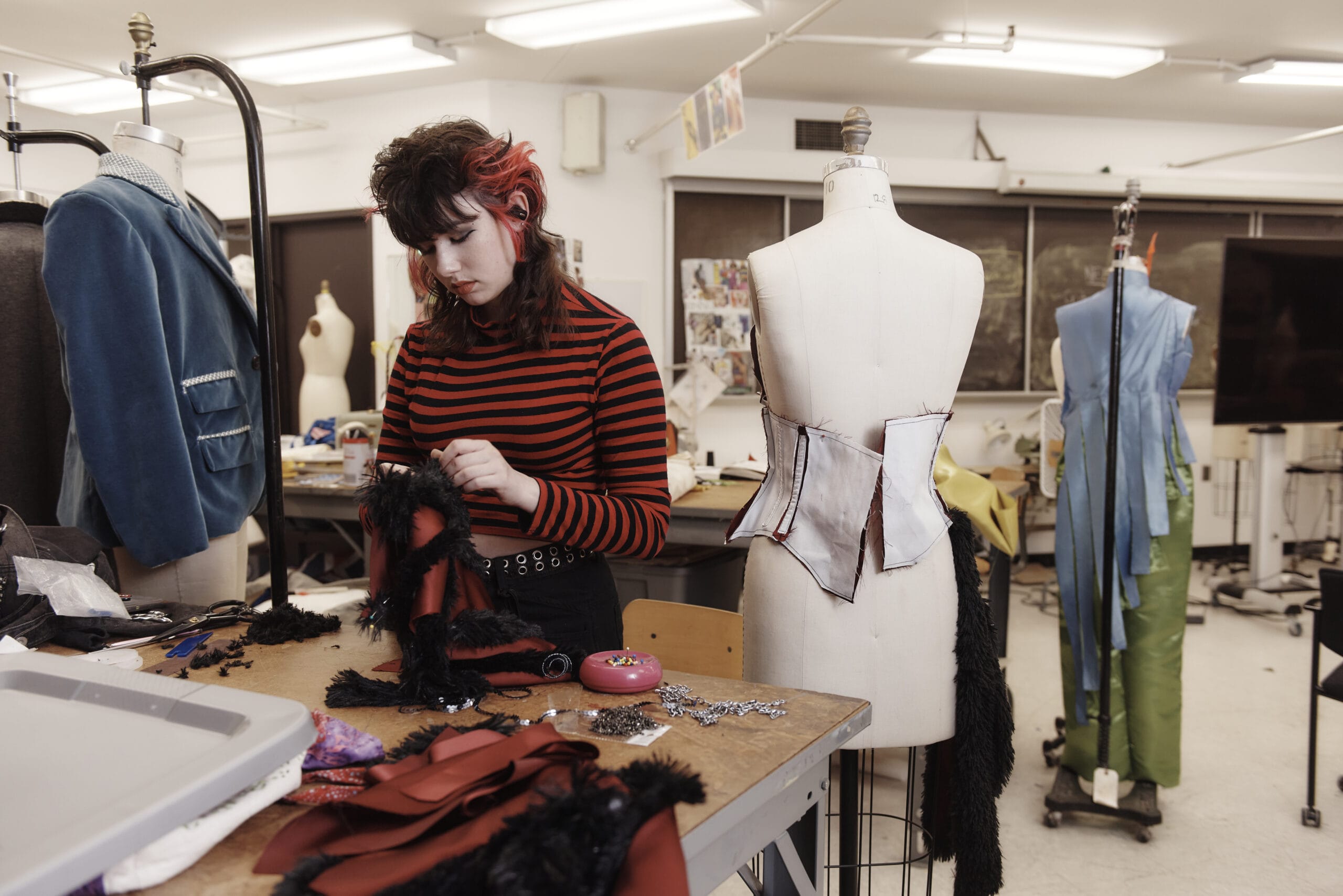 A person in a striped shirt works on a garment at a table cluttered with sewing materials, embodying the spirit of MassArts fashion design courses. Surrounding mannequins display partially completed clothing, while the room boasts sewing equipment and patterns on the wall.