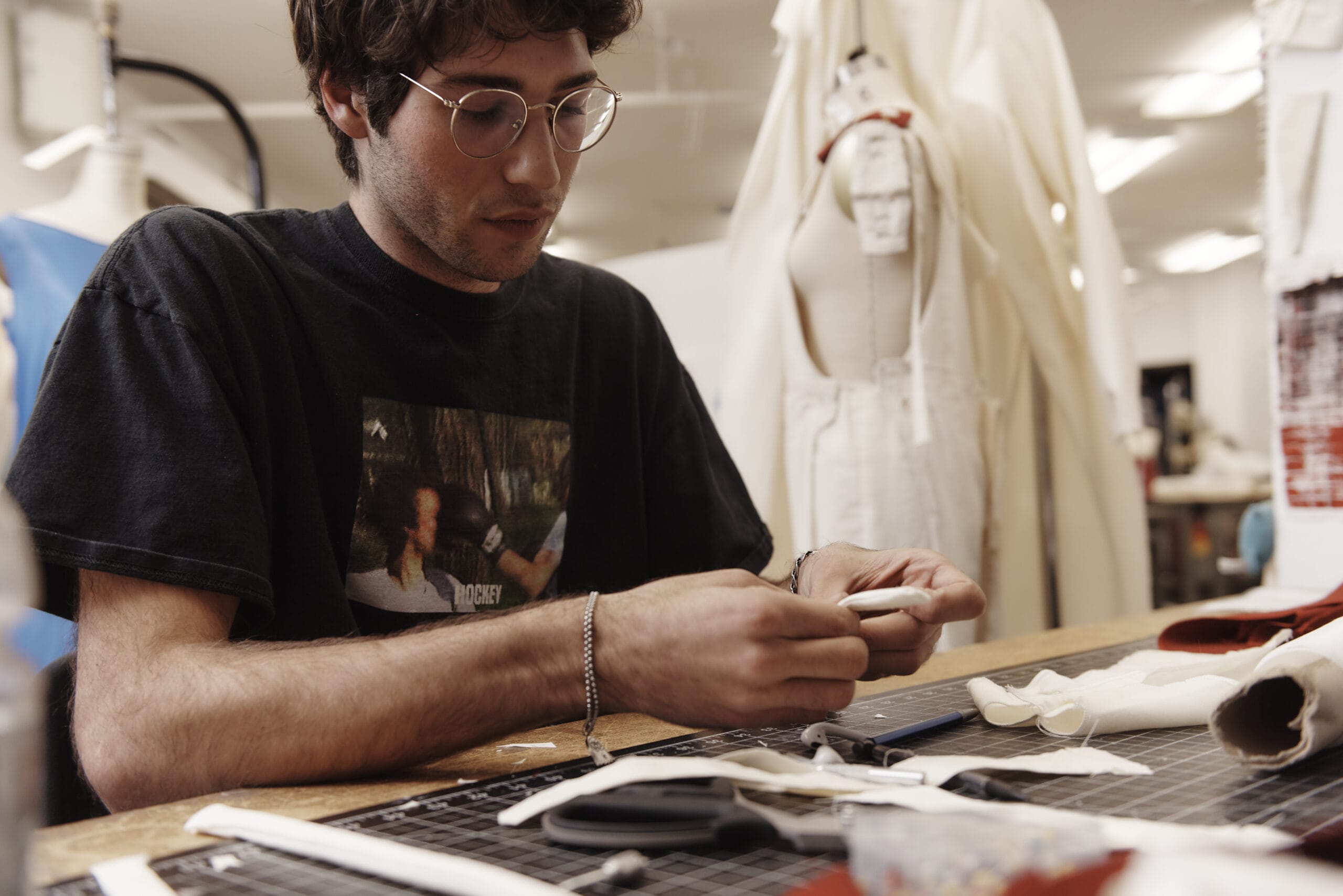 A person in glasses and a graphic t-shirt works diligently at a table with sewing materials in a workshop, embodying the creative spirit of the MassArt Fashion Design program. Scissors, fabric strips, and a mannequin showcasing a white garment are visible in the background.