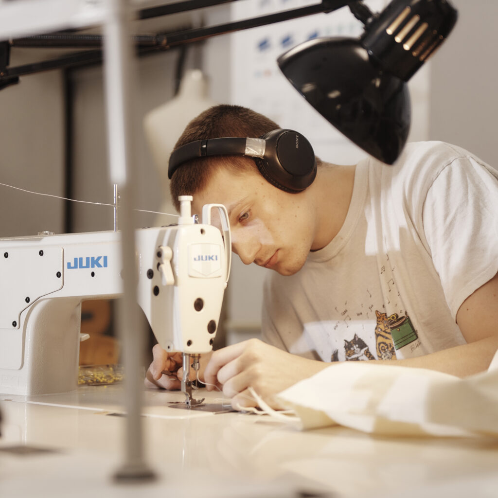 A person wearing headphones is focused on sewing fabric with a Juki sewing machine in a well-lit workspace, crafting pieces for their MassArt Fashion Design portfolio. A lamp shines on the area, highlighting their concentration.