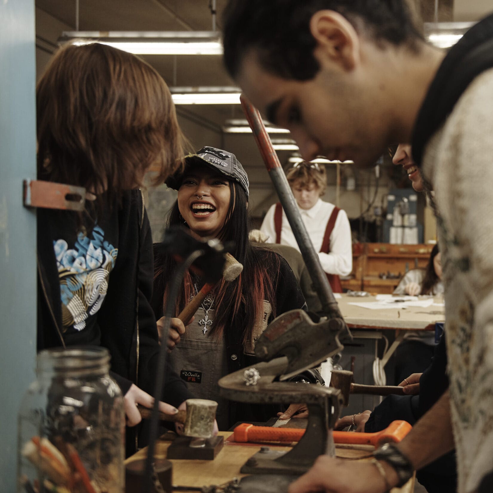A diverse group of young people are gathered in a lively workshop. They are engaging in a jewelry crafting activity, using tools and materials on a wooden table. Everyone appears focused, with one person smiling and laughing, adding to the collaborative atmosphere.