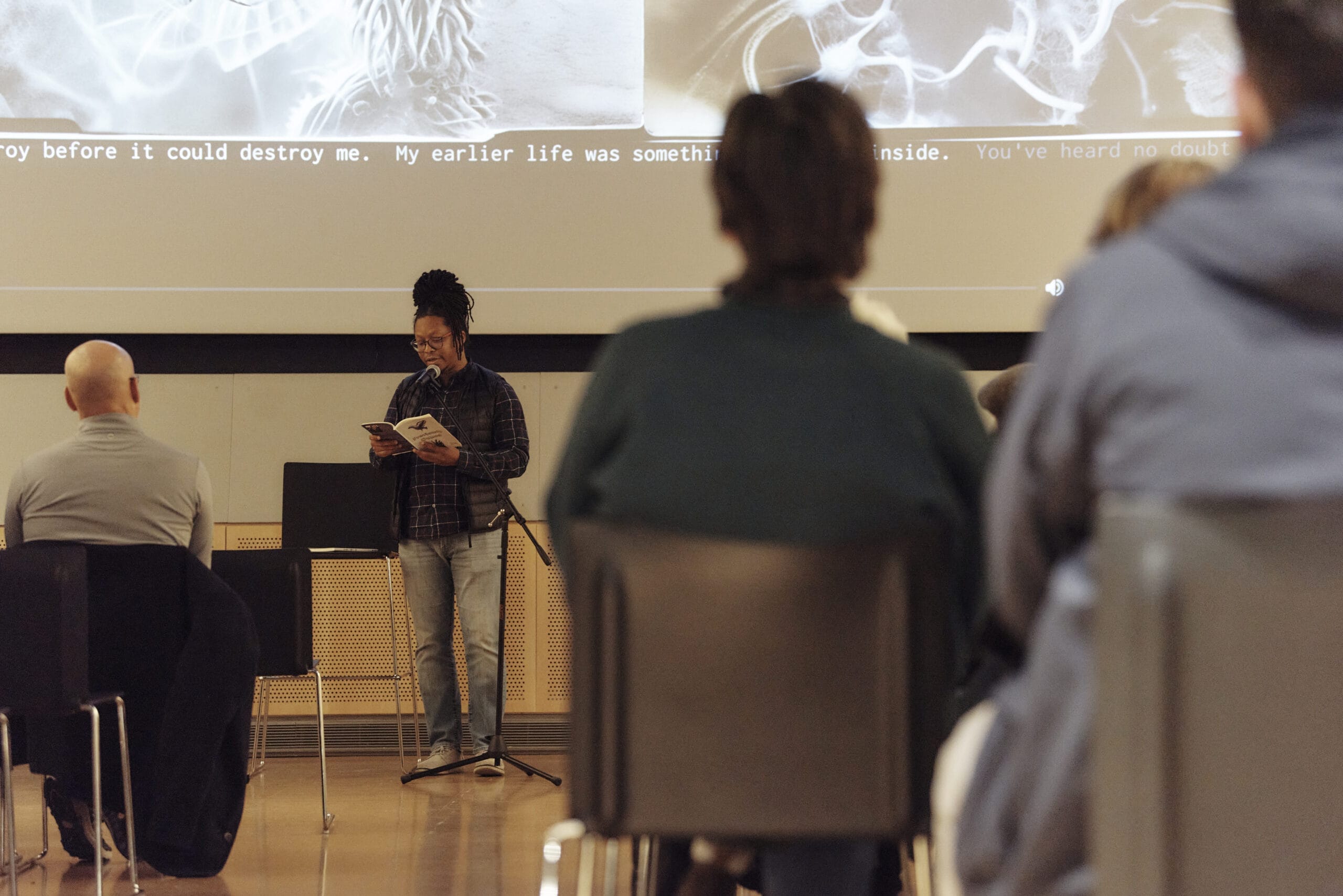 A person stands on stage, book in hand, engaging in creative writing. A microphone captures each word as the seated audience members eagerly listen. On the wall, a projection displays text in a dimly lit room, adding to the atmosphere of literary exploration.