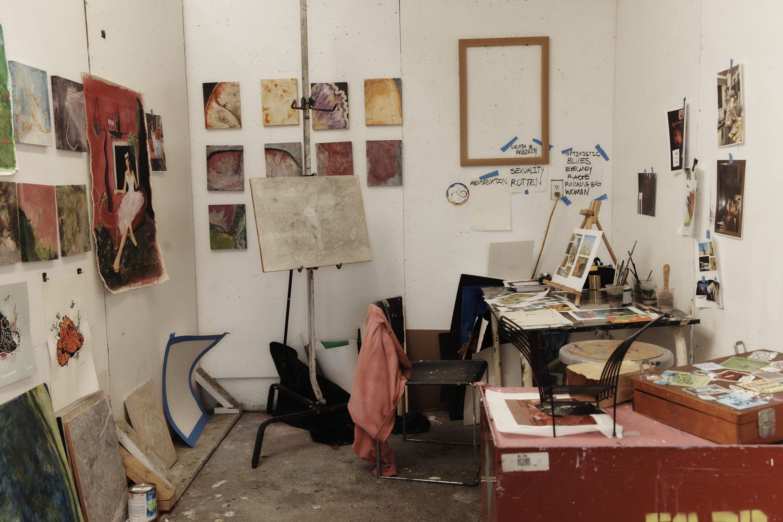 An artist’s studio with various paintings on the walls, an easel with a blank canvas, and a cluttered desk with art supplies. A pink coat hangs on a chair. The room has a creative, unfinished atmosphere.
