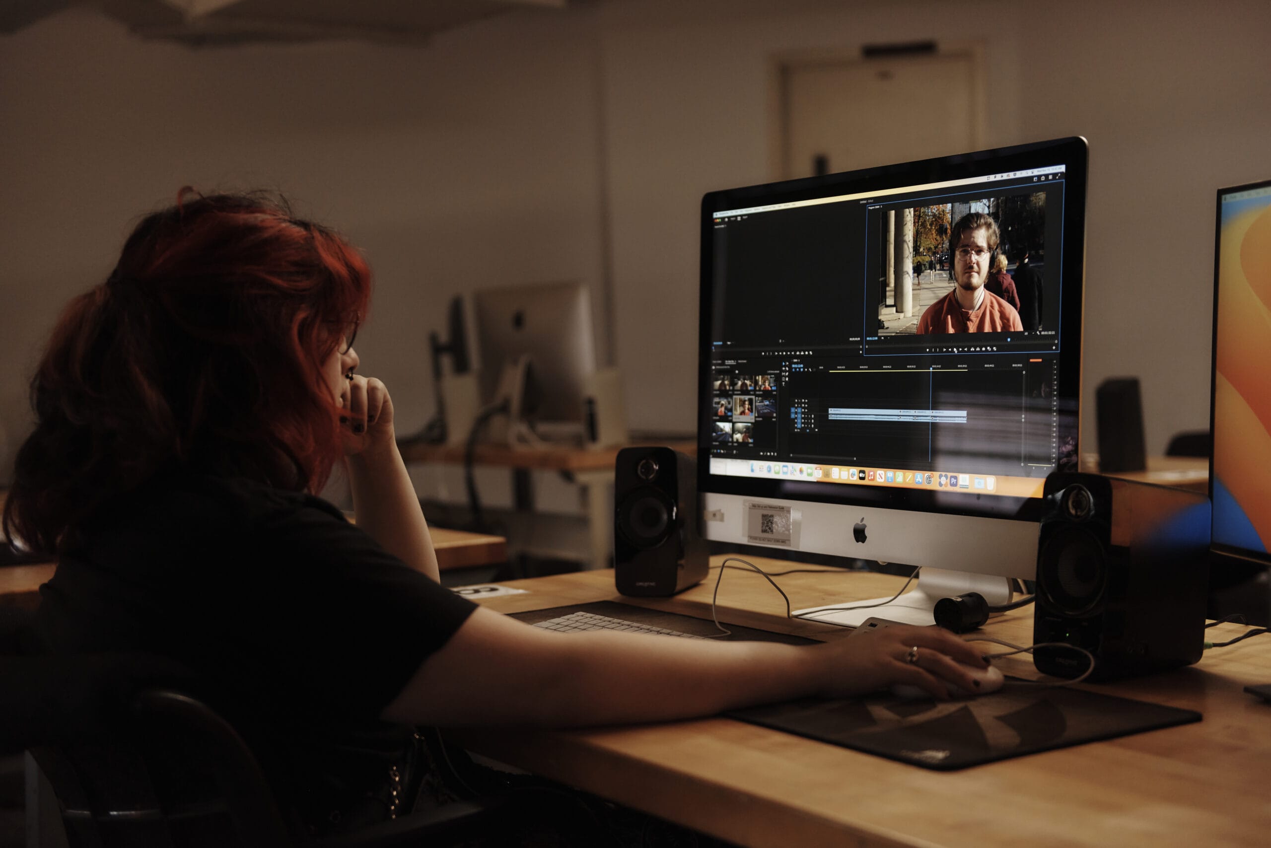 A person with long hair meticulously edits footage on video editing software, likely part of a top-tier film and video production program. The screen shows a video timeline and a man in frame, surrounded by speakers and a monitor in an office or studio setting.