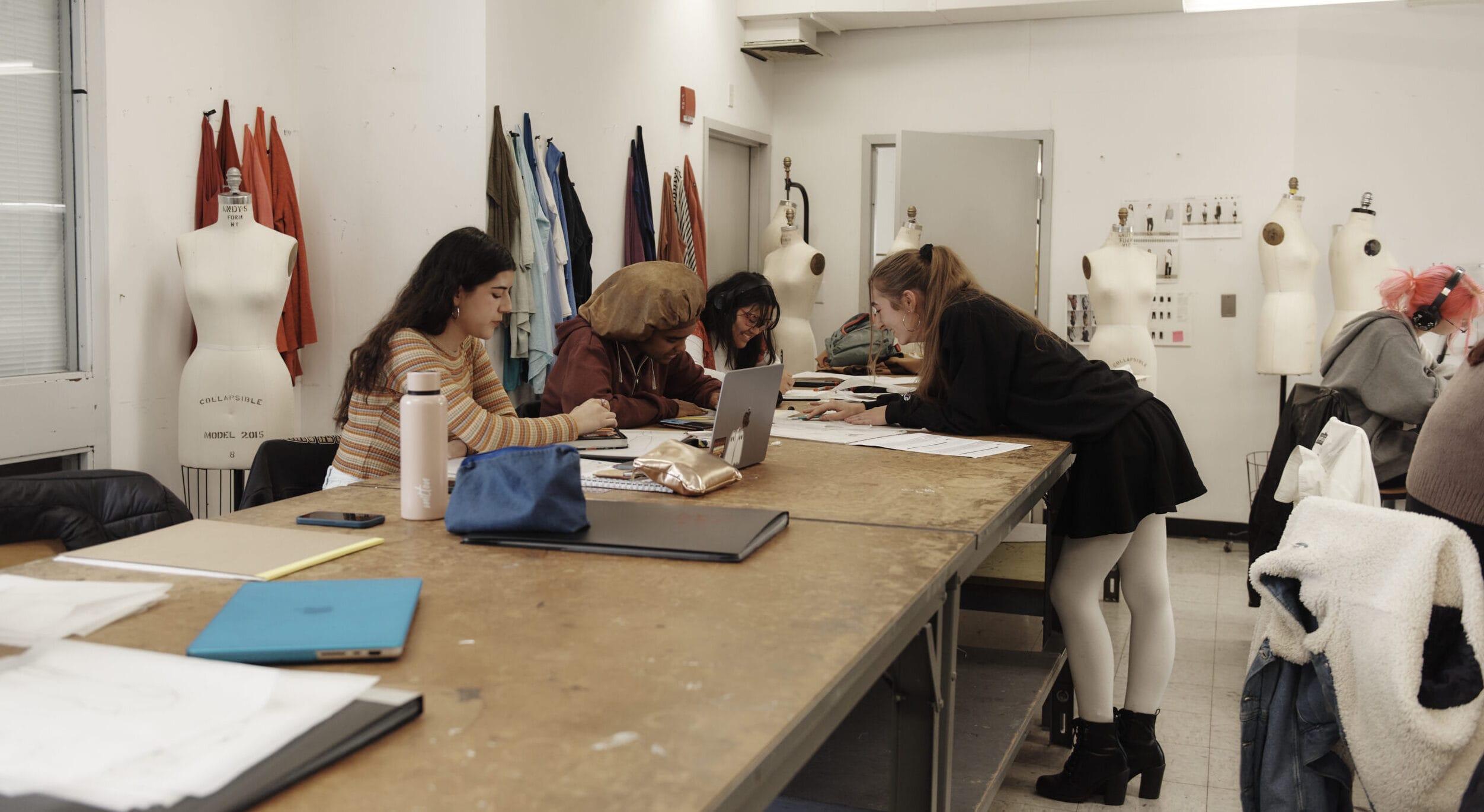 The vibrant MassArt Fashion Design studio buzzes with creativity, as several people engage in work. Mannequins draped with fabric line the walls, while sketchbooks and laptops cover the table. This collaborative atmosphere is a testament to the dynamic energy of MassArt fashion design courses.