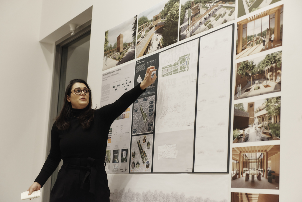 A woman in a black outfit and glasses gestures toward architectural design posters on a wall, showcasing building plans, renderings, and photographs. She seems to be explaining the designs, possibly highlighting various careers in architecture, in an indoor setting.