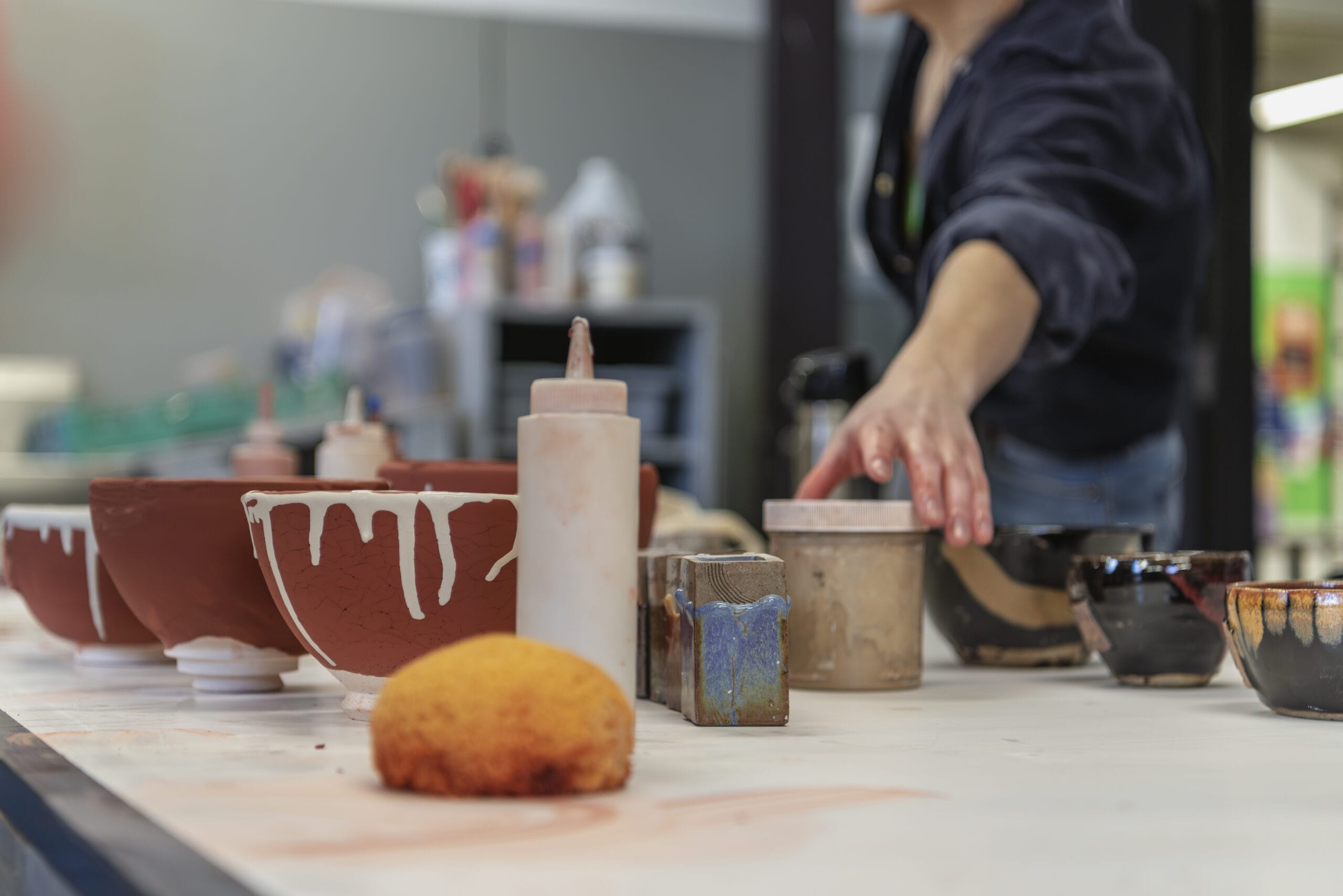 A person with a BFA reaches for a small container on a table filled with pottery supplies. Various glazed and unglazed ceramic bowls are visible, along with a sponge and bottles for glaze. The background reveals the vibrant hustle of their ceramics workshop.