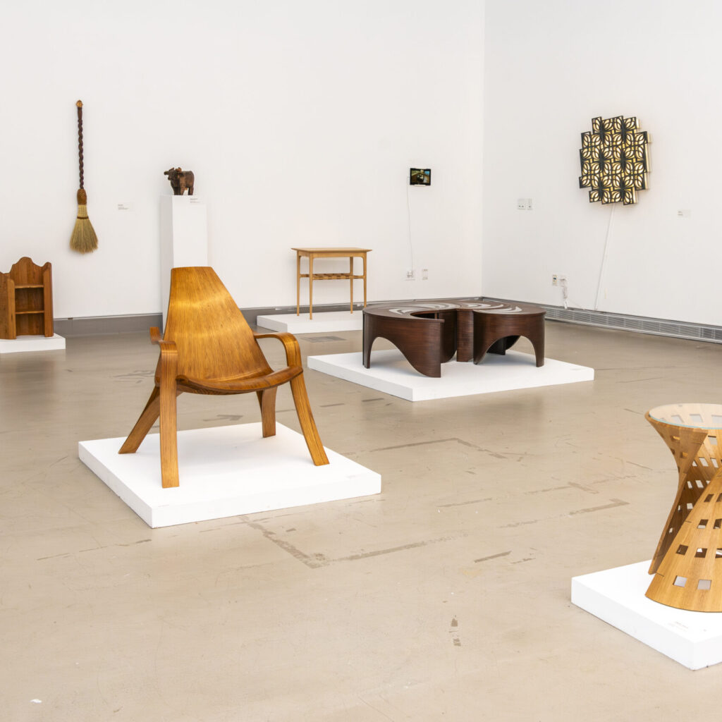 A gallery room showcasing exquisite furniture design, featuring wooden pieces like chairs, a broom, a sculpture, and a table. Each item rests on a white platform, while small artworks adorn the walls against the backdrop of a light tan floor.