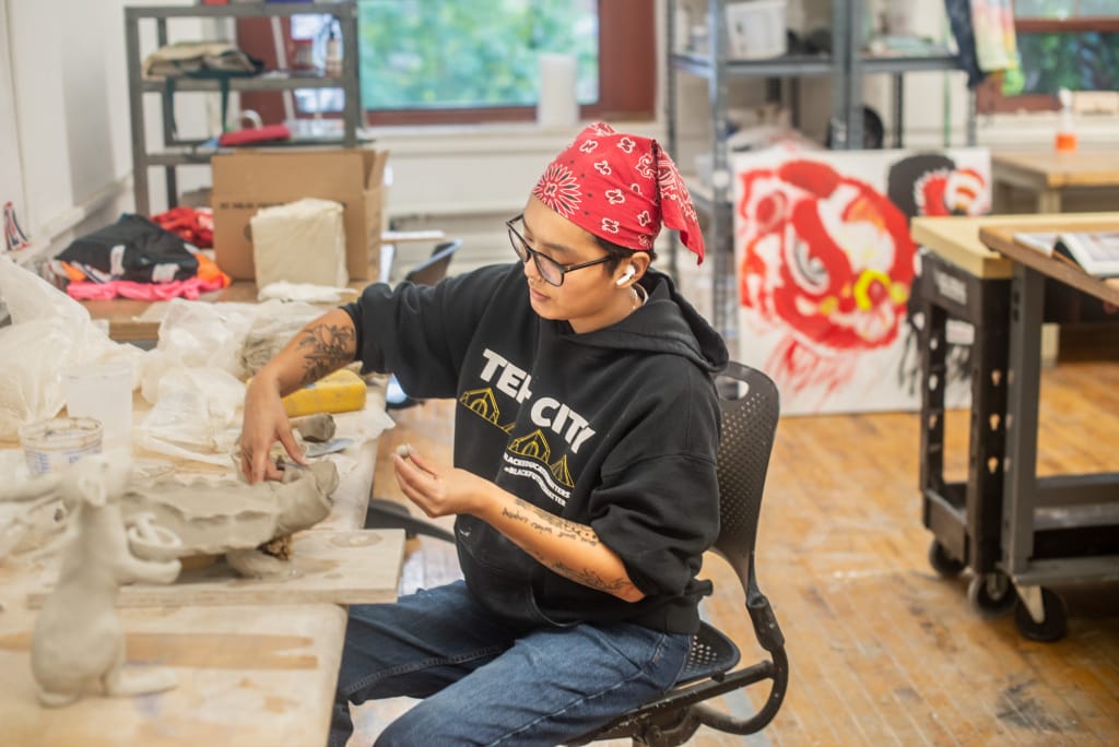 A person with glasses and a red bandana sculpts clay at a wooden table in an art studio. They wear a black hoodie and jeans. The room has art supplies, a window, and a colorful painting on the wall.