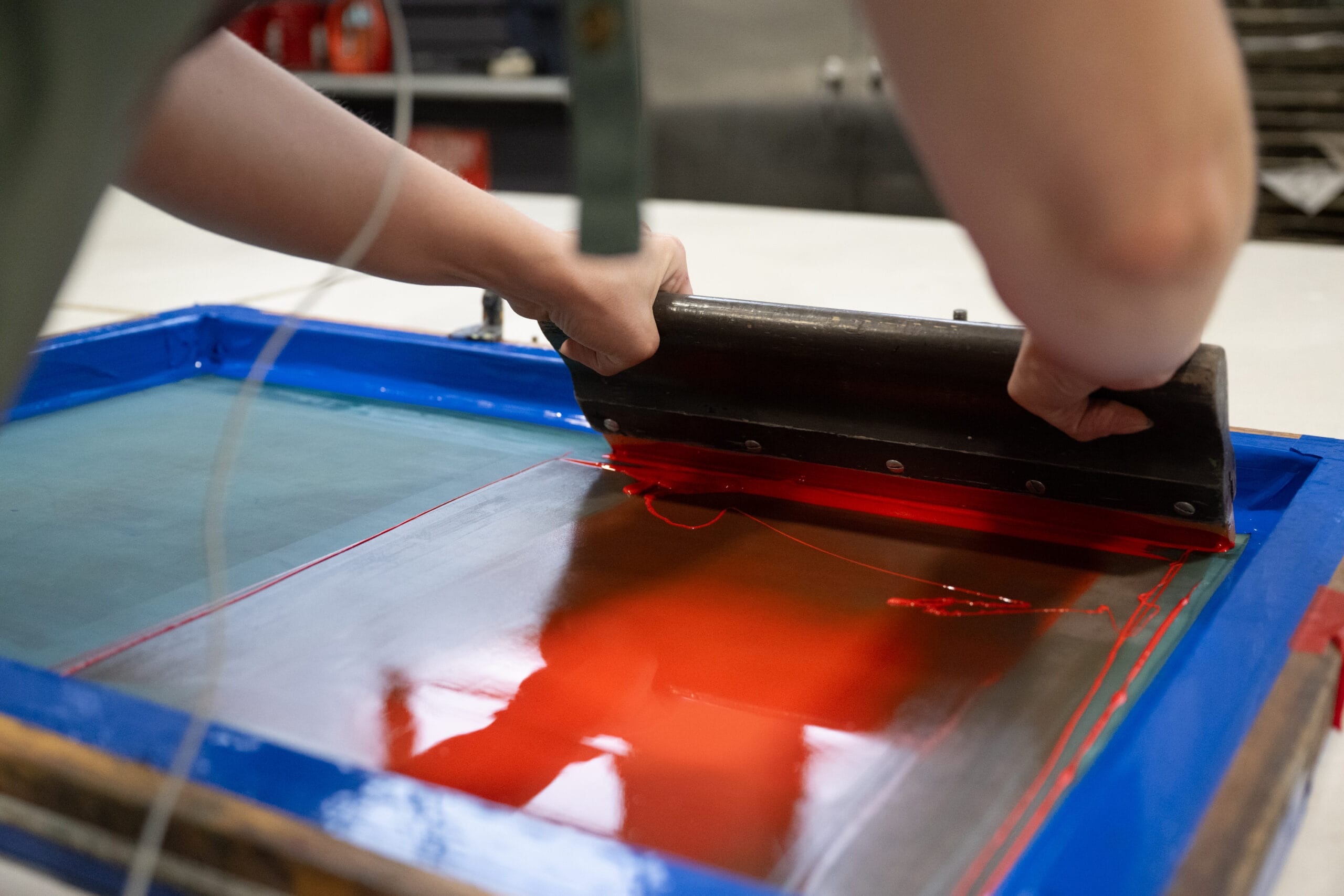 A person is screen printing with a squeegee, spreading bright red ink over a framed screen. The work is taking place on a flat surface, with the ink and screen partially covering the area.