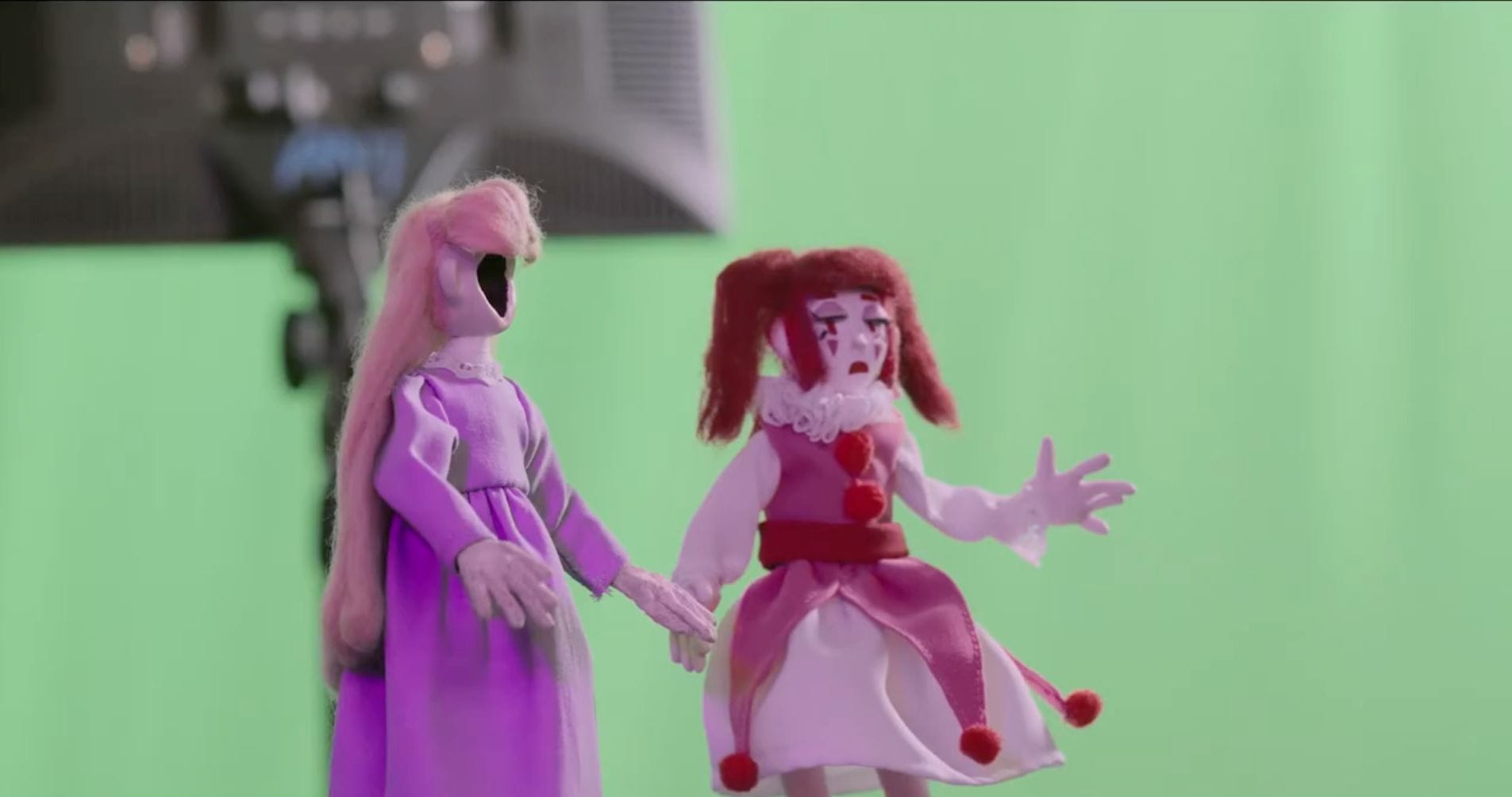 Two puppets with no facial features stand in front of a green screen background. The puppet on the left, perfect for a major in animation project, has long pink hair and a purple dress. Meanwhile, the other sports a pink and red clown costume with red pigtails, alongside visible studio lighting.