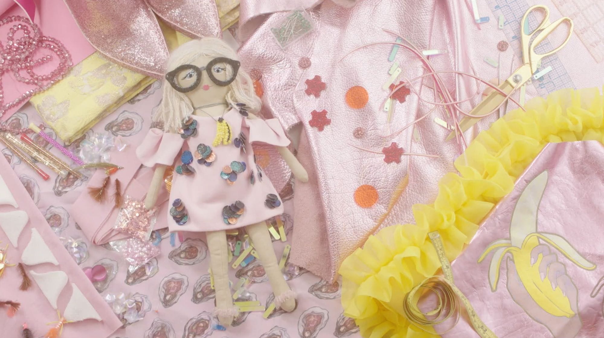 Video still of a blond handmade doll laying on pink fabric