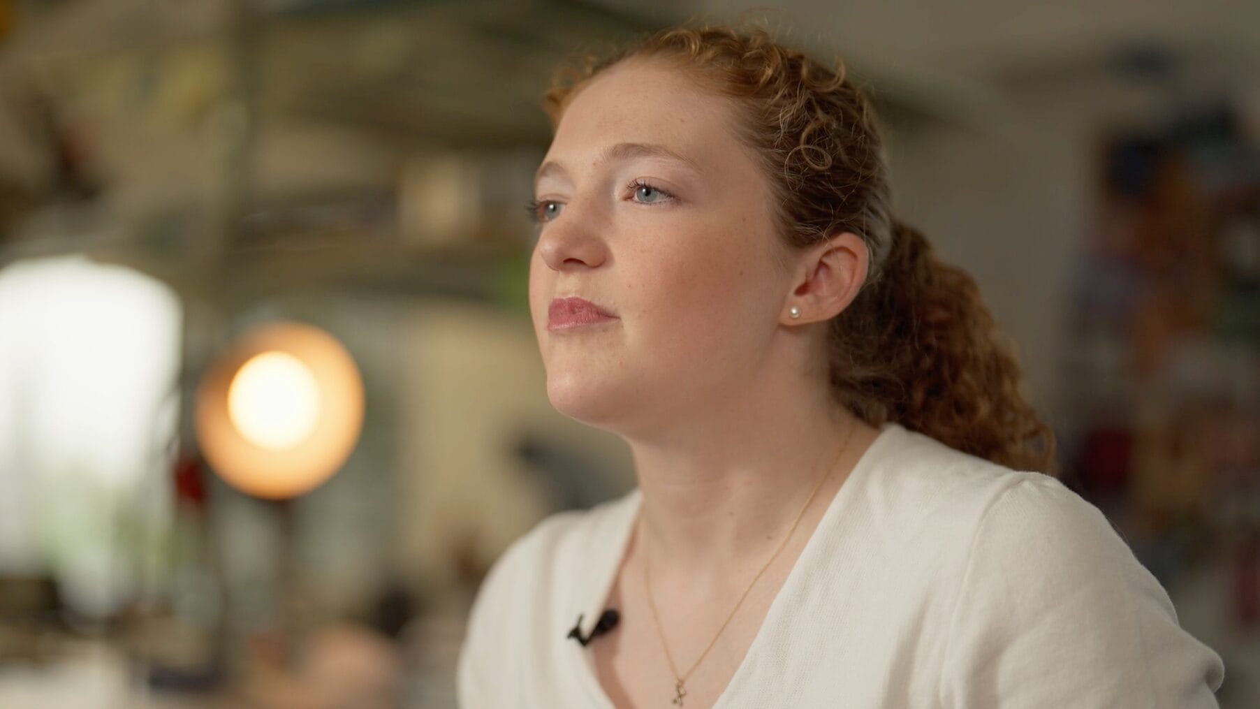 A young woman with curly red hair and fair skin, wearing a white top and a small necklace, looks thoughtfully into the distance in a softly lit indoor setting that hints at modern industrial design elements.