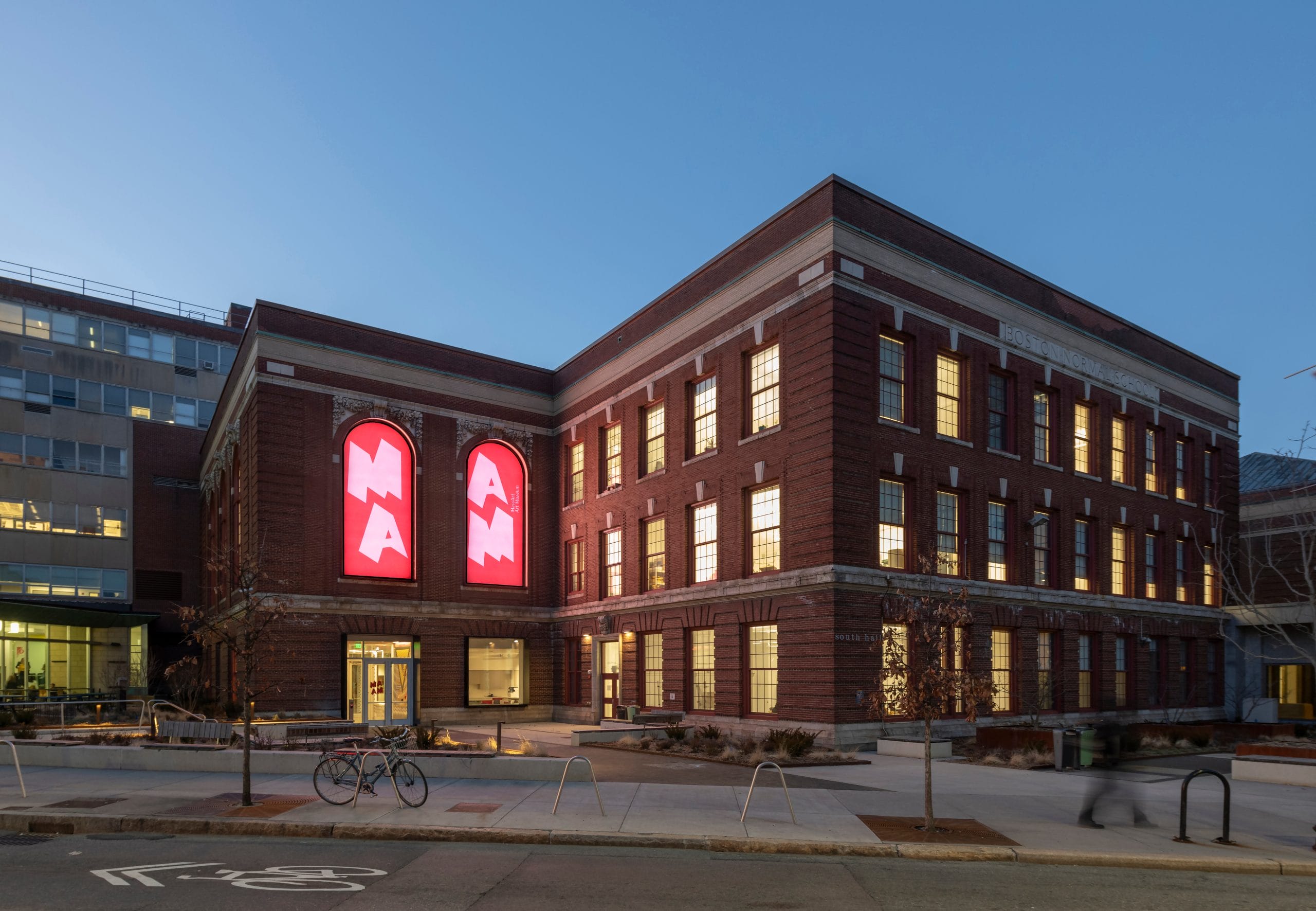 Exterior of a brick building at dusk with large red illuminated signs displaying "MAAM". The building has numerous windows, some lit from within.