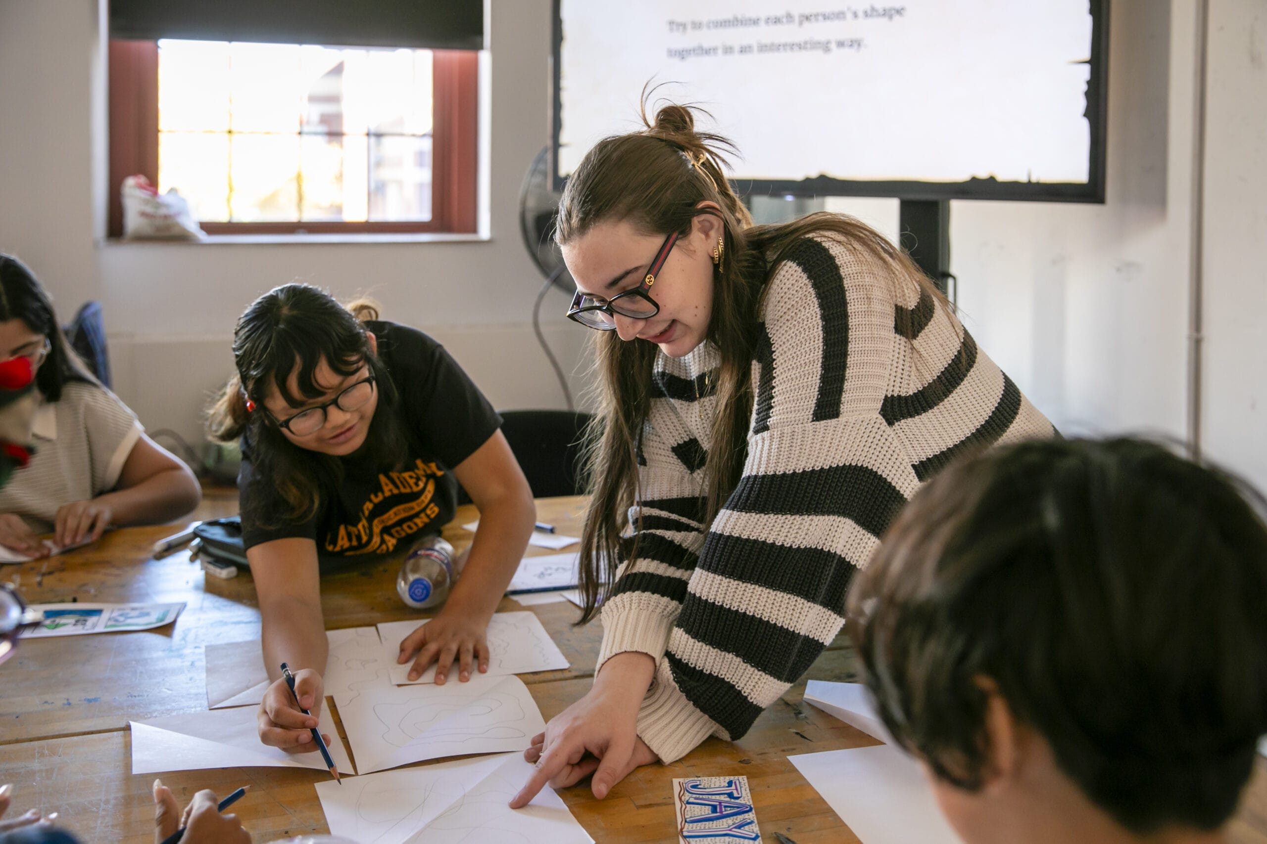 A group of students is gathered around a table, immersed in an art education project. A woman in a striped sweater leans over, pointing at one of the drawings. Papers and art supplies are scattered across the wooden table, fueling their creative collaboration.