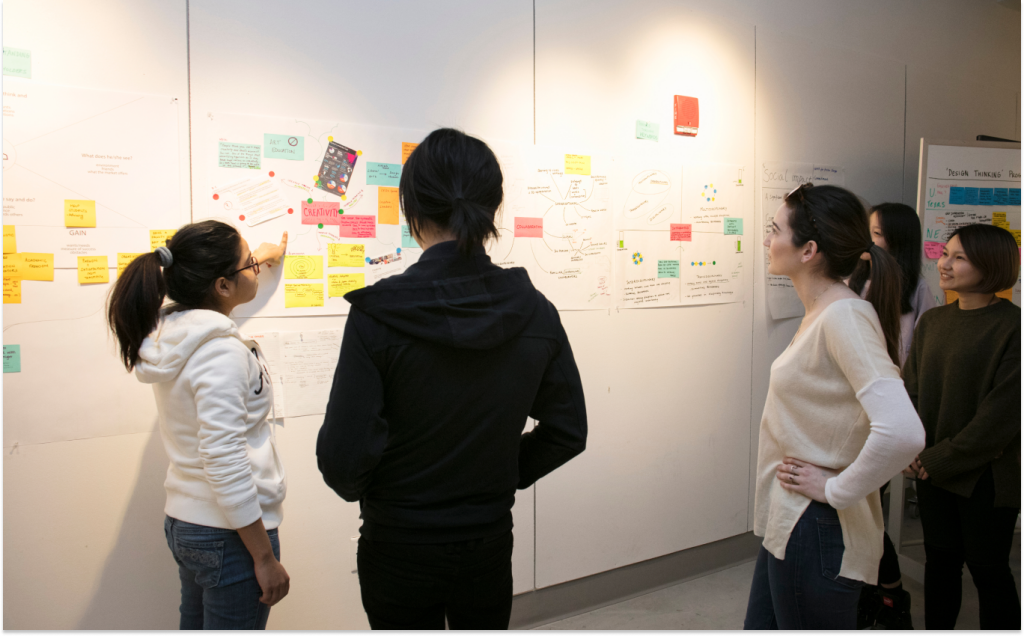 A group of five people stands in front of a wall brimming with innovation, featuring various papers and colorful sticky notes. One person points at the design ideas while others listen and observe attentively. They are dressed casually in a well-lit room.