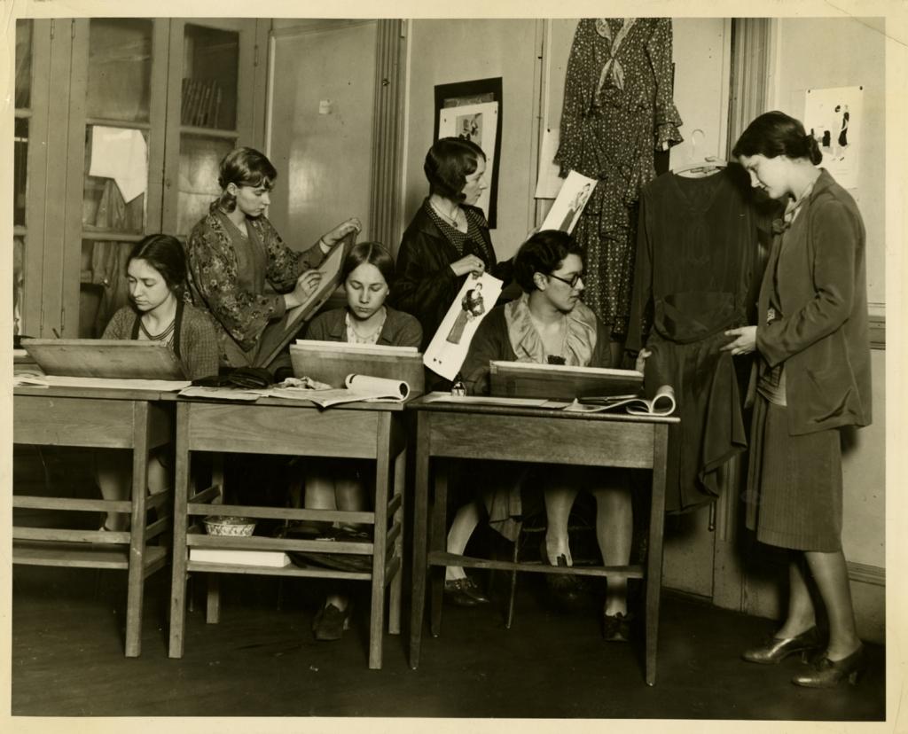 A black-and-white photo of six women in a classroom at ºìÁì½í¹Ï±¨ Fashion Design. Three are seated at desks with drawing materials, while three stand behind, examining dresses and sketches. The room features wooden furniture and dress patterns displayed on the walls, capturing a creative atmosphere.