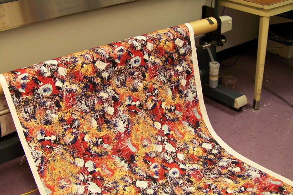 A large printer outputs a colorful, abstract fabric design with a blend of red, orange, black, and white patterns. Situated in a room with a purple floor and nearby table holding a computer and sanitizer, this setting resonates with the innovative spirit of MassArt Fashion Design.