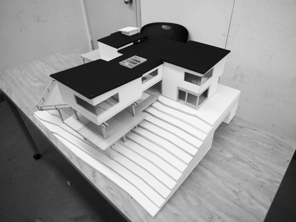 A black-and-white image of a modern architectural model on a table captures the essence of a recreational center. The design features multiple levels and flat roofs, reminiscent of structures near Jamaica Pond. This intricate paper model sits near a chair against a plain wall.