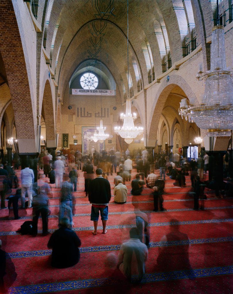 The transnational interior of a large mosque features a rich red carpet, graceful chandeliers, and arched brick ceilings. Blurred figures sit or walk beneath walls adorned with banners and calligraphy, as light streams through the upper windows.