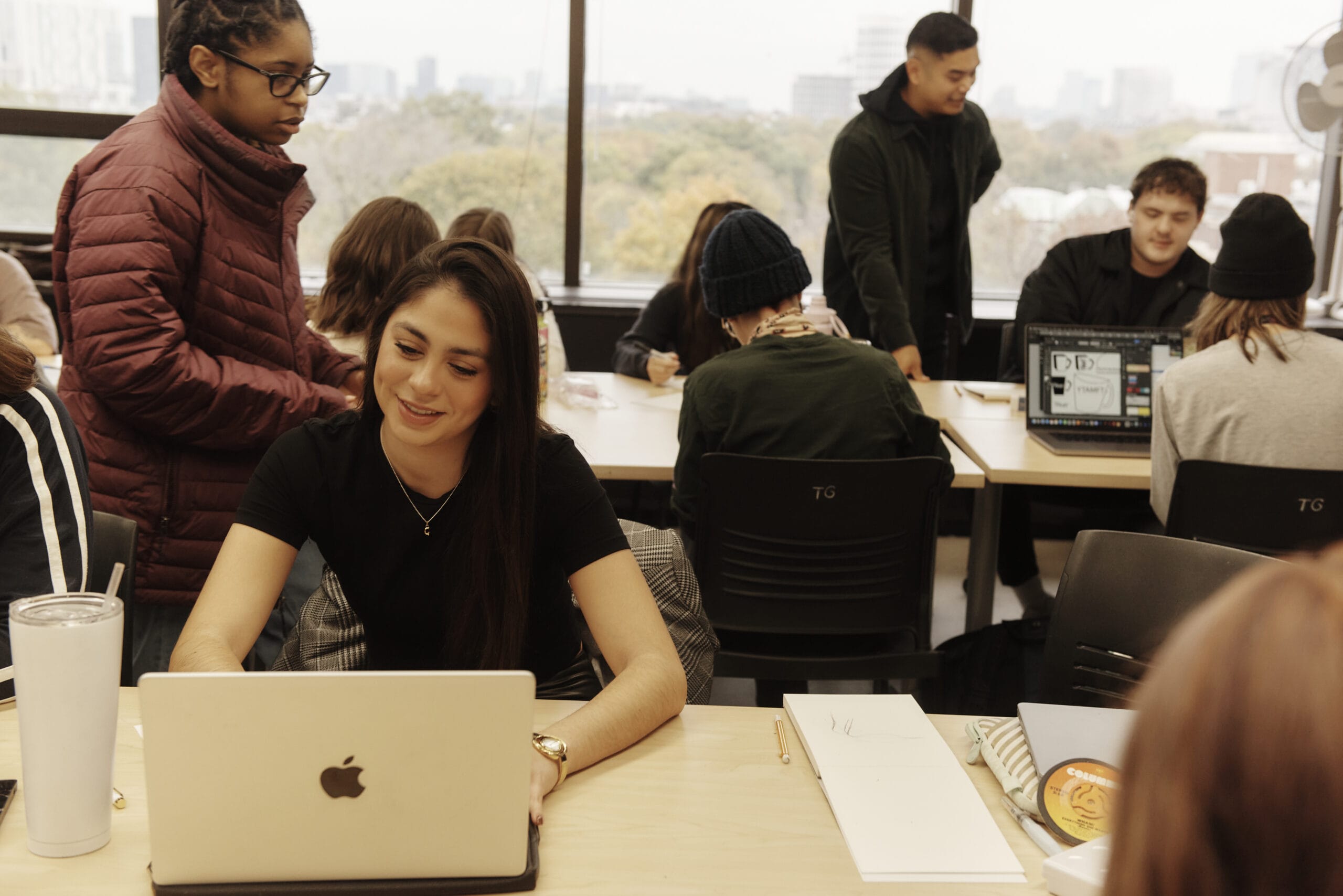 In an atmosphere buzzing with collaboration, a classroom thrives as students engage dynamically in activities. One smiles at their laptop, embodying institutional effectiveness, while others gather in groups near a window framing an overcast sky, reflecting thoughtful conversations and teamwork.