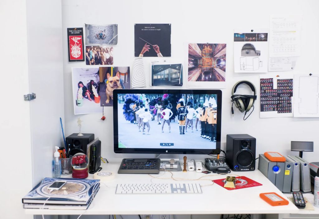 A cluttered desk showcases a computer displaying a vibrant parade scene. The wall behind brims with photos, posters, and headphones. Among the books, keyboard, speakers, and various electronic devices lies a self-service hub of creativity and inspiration.