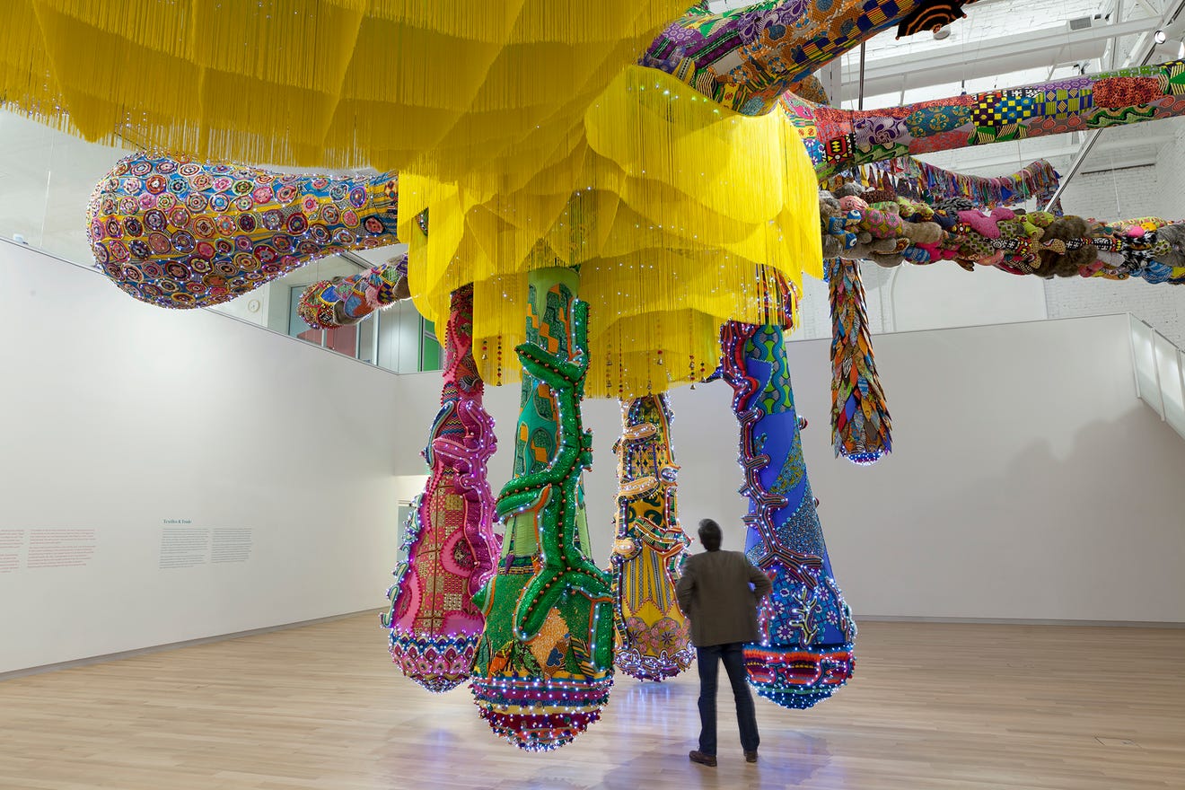 A man observes a large, colorful art installation by Joana Vasconcelos, featuring intricate bead-like patterns and long, dangling yellow fringe. Suspended from the ceiling in MassArt Art Museums spacious gallery, it complements the light wooden floors and white walls beautifully.