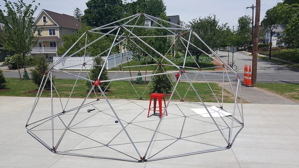 A large 2V geodesic dome frame made of interconnected metal rods stands on a concrete surface in a suburban area. A single red stool is placed inside the dome. Houses, trees, and road construction signs are visible in the background.