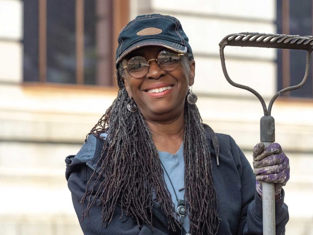 Smiling woman in sunglasses and a denim cap holds a gardening rake at the Garden for Boston. She wears a blue jacket, gloves, and has long gray braided hair. In the background, near the MFA entrance, theres a building with large windows enhancing the scene crafted by local artists.