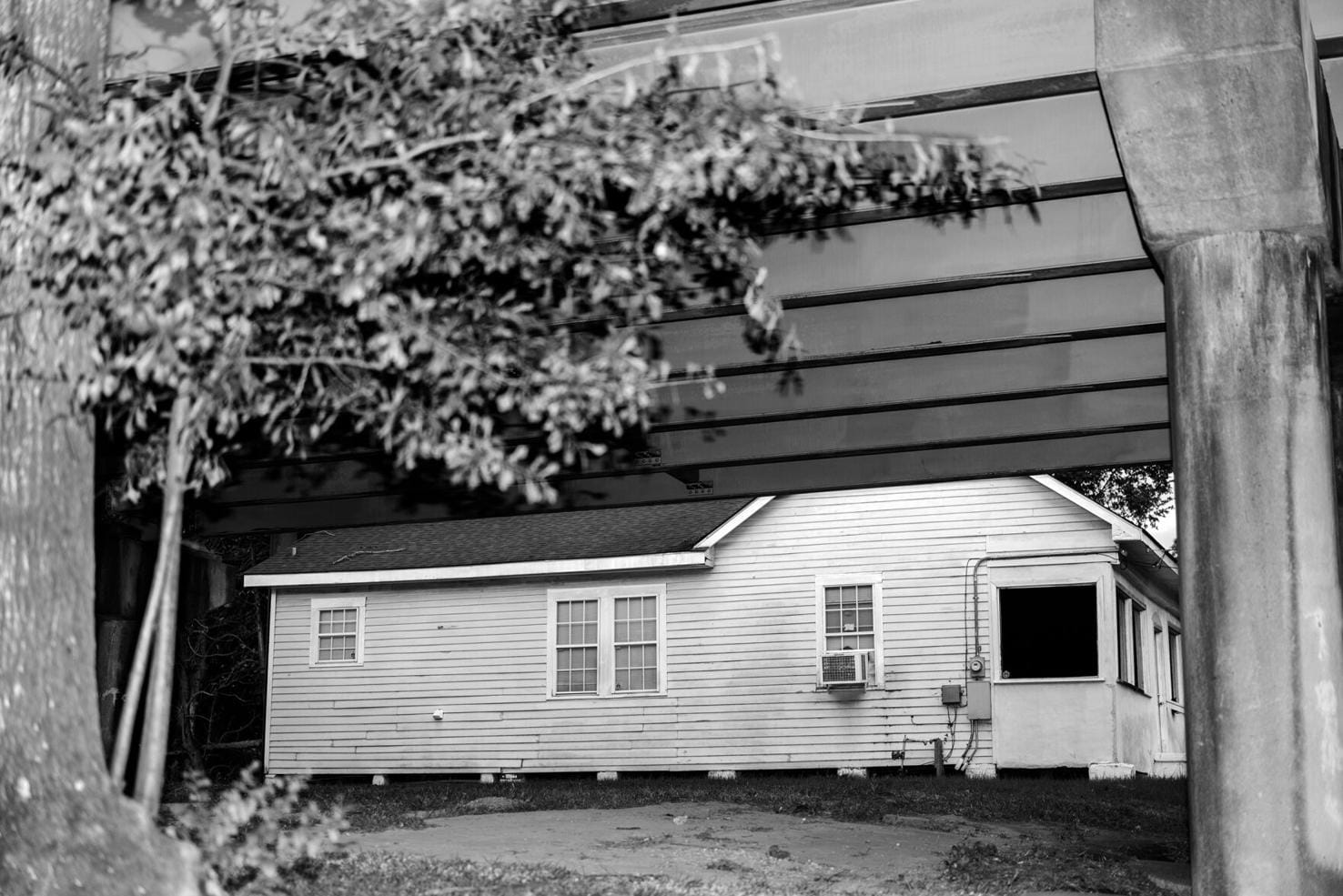A small, white wooden house with multiple windows and an air conditioning unit is partially obscured by a large overhead bridge in Old South Baton Rouge. Tree branches frame the left side of the image, capturing a slice of history reminiscent of displays at Capitol Park Museum. The scene is in black and white.
