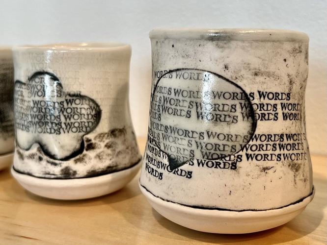 Ceramic cups with an irregular shape feature repeated text WORDS forming abstract patterns on their surfaces, perfect for a cup of coffee. The background is a smooth, light-colored finish with subtle dark accents, displayed as if at the Glassell Gallery on a wooden surface.