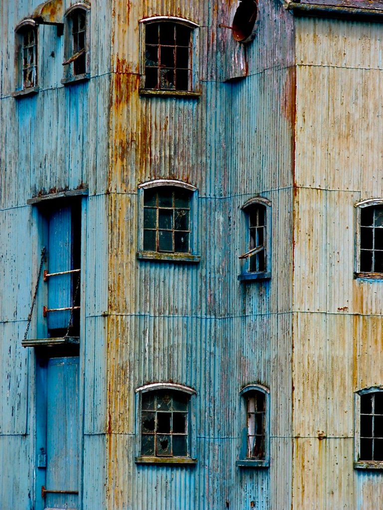 An old, weathered building facade at the abandoned docks, clad in vertical corrugated metal siding with faded blue and rust hues. It features several small arched windows with rusty frames and a somber blue door.
