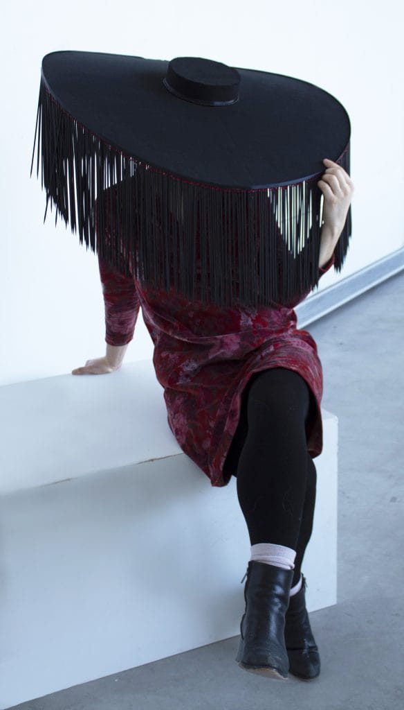 A person wearing a large, wide-brimmed black hat with fringe sits on a white block. They wear a red patterned dress, black leggings, and black boots, while holding the hat with one hand. The background is plain white.