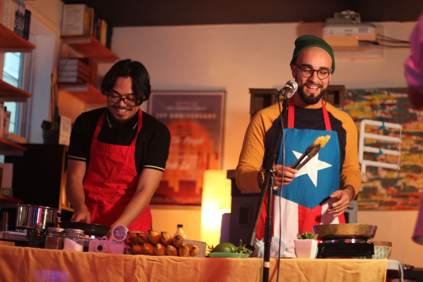 Two people are cooking at a table, both wearing aprons. The person on the right holds tongs and wears an apron with a flag design. Various cooking utensils and ingredients, including fish sauce for their adobo dish, are on the table. Theres a colorful background and a microphone in view.