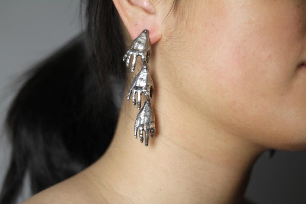 A person wearing an earring featuring a series of small metallic hands hanging in a row. The background is neutral, and the person has dark hair tied back.