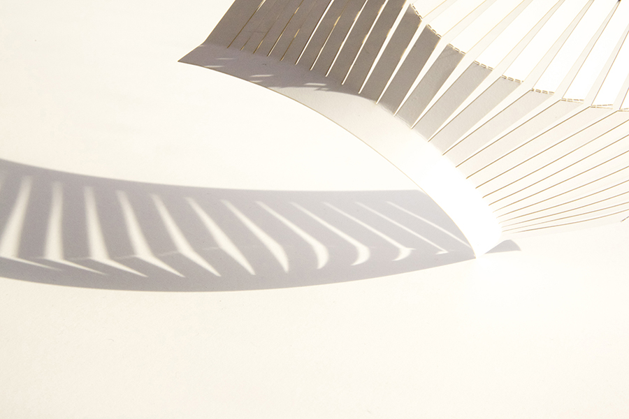A spiral pattern of white slats casting shadows on a smooth, pale surface. The composition creates a dynamic, abstract play of light and shadow, forming a visually striking curved design.