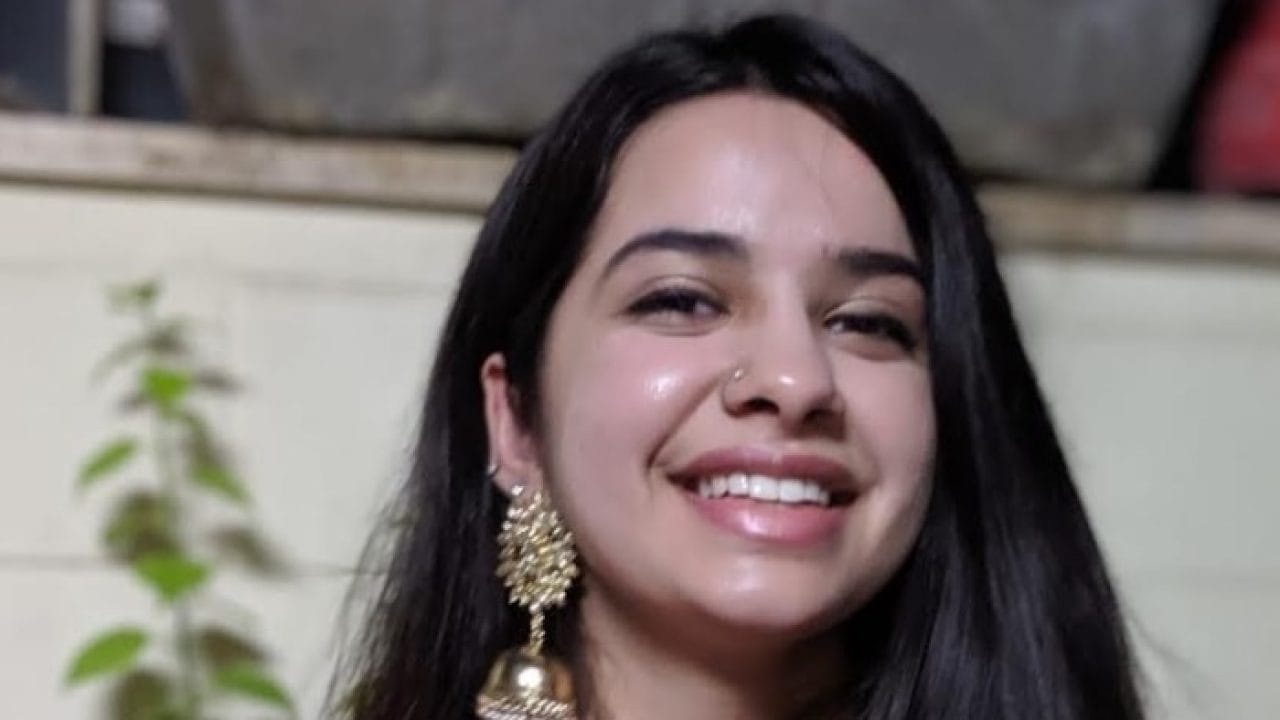 Smiling Anushka Suji, with her long dark hair and gold earrings, stands in front of a light-colored wall. Green plants form a serene backdrop for the Studio Designer from the Creative Department.