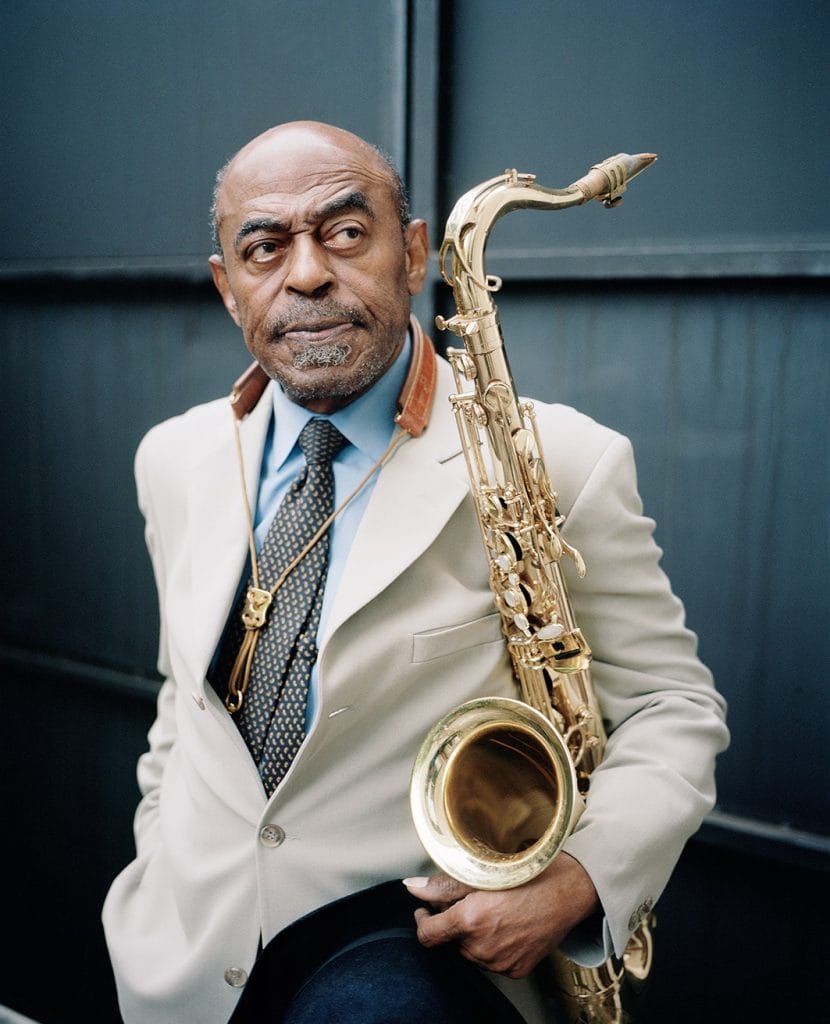 An older man with a bald head and mustache, reminiscent of Archie Shepp, holds a saxophone. Clad in a beige suit jacket, blue shirt, and patterned tie, he stands against a dark, industrial backdrop, gazing thoughtfully to the side.