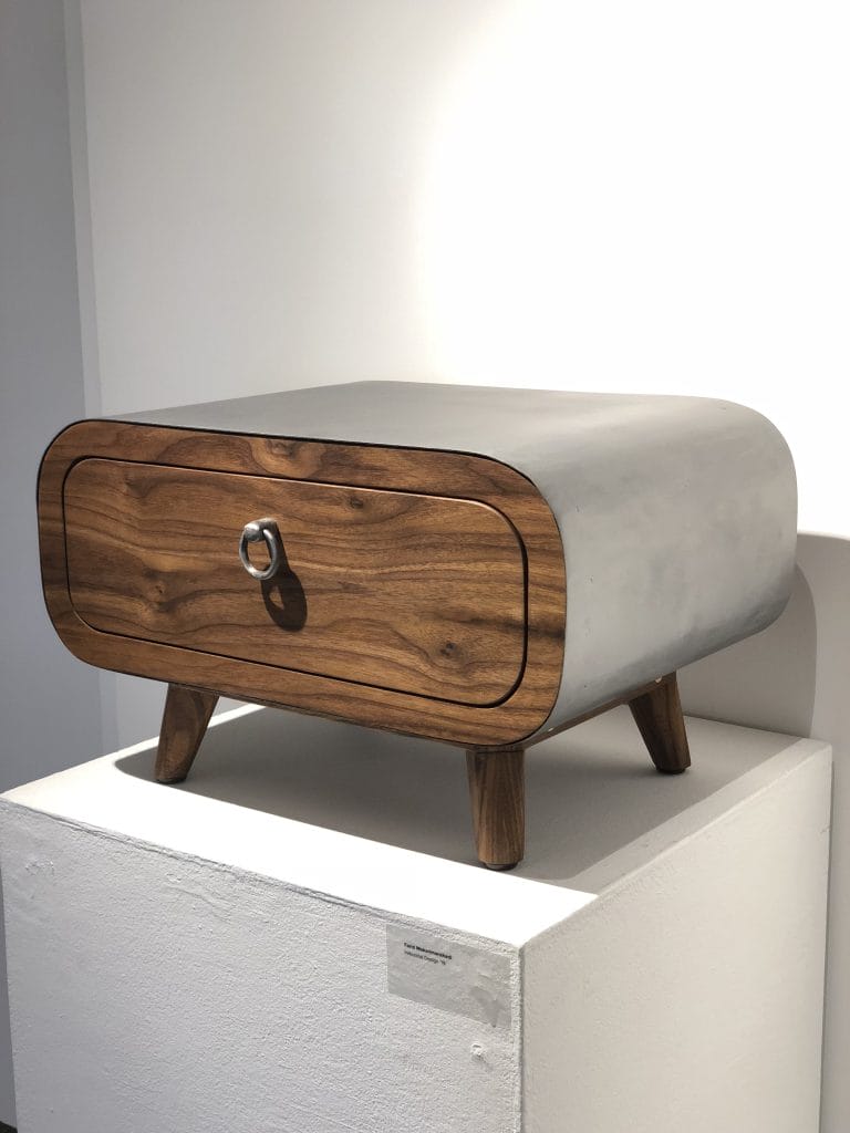 A modern sidebed table with a rounded concrete top and wooden front, featuring a single drawer with a circular handle. It stands on four angled wooden legs and sits on a white pedestal in a well-lit room.