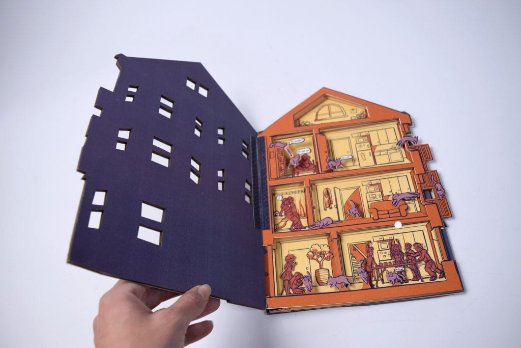 A hand holds a pop-up book titled Curious House, shaped like a home. The left page is black with window cutouts, while the right reveals an orange house interior, showcasing multiple floors and rooms filled with detailed scenes and characters.