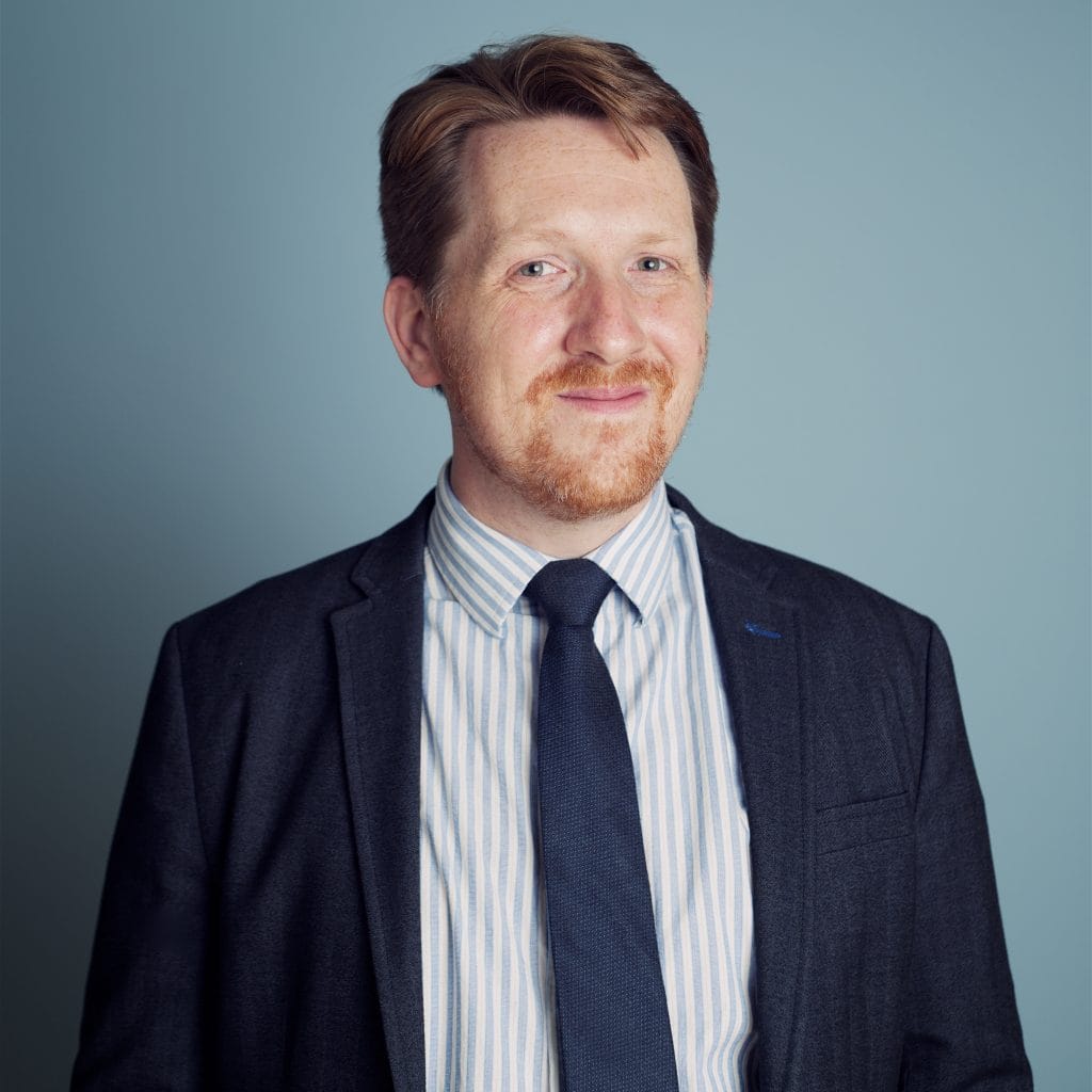 A man with short red hair, a beard, and a mustache is smiling. He is wearing a dark suit jacket, a white and blue striped shirt, and a dark tie. The background is a solid light blue color.